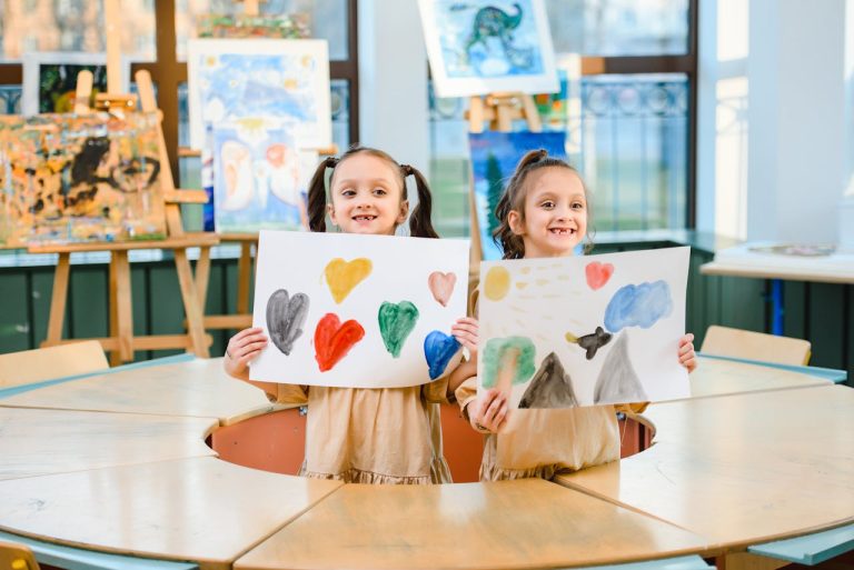 Two children showcasing colorful paintings in an art classroom setting.